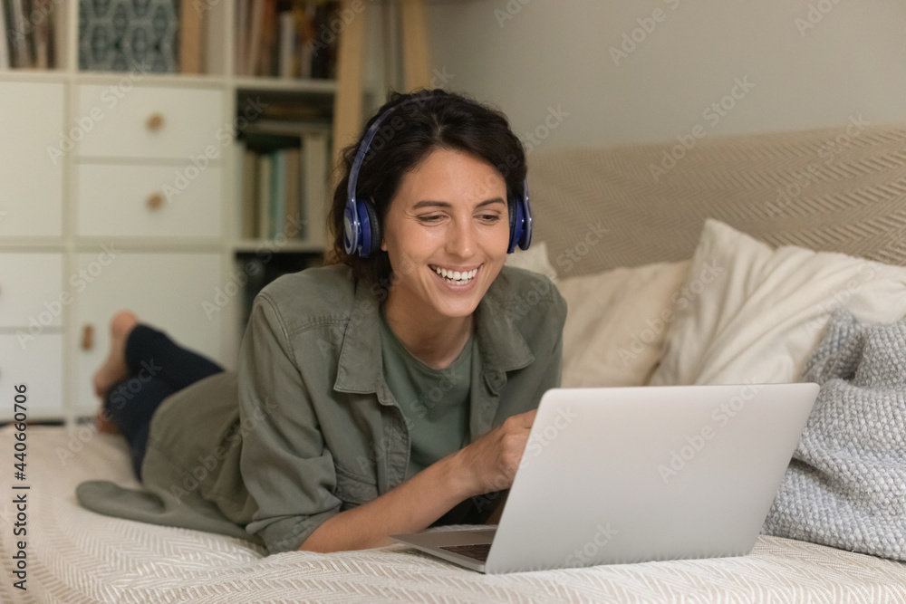 Excited woman in wireless headphones using laptop, lying on couch, looking at laptop screen, watching movie, having fun with device, involved in internet meeting, chatting online with friends Excited woman in wireless headphones using laptop, lying on couch, looking at laptop screen, watching movie, having fun with device, involved in internet meeting, chatting online with friends