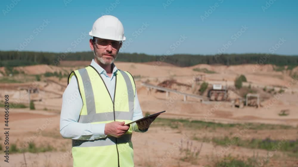 Handsome male sand quarry worker wearing safety uniform goggles and hard hat working with digital tablet computer. Modern engineer looking at camera. Sand mining project professional on warm sunny day Handsome male sand quarry worker wearing safety uniform goggles and hard hat working with digital tablet computer. Modern engineer looking at camera. Sand mining project professional on warm sunny day