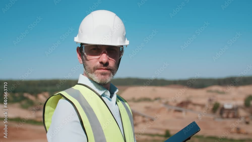 Portrait of handsome male sand quarry worker wearing safety uniform goggles and hard hat holding digital tablet. Modern engineer looking at camera. Sand mining project professional on warm sunny day Portrait of handsome male sand quarry worker wearing safety uniform goggles and hard hat holding digital tablet. Modern engineer looking at camera. Sand mining project professional on warm sunny day