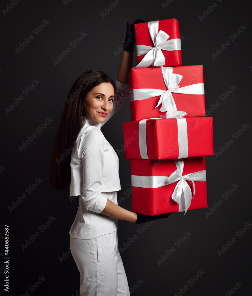 Attractive young positive woman doctor dentist in white uniform and latex gloves standing and holding heap of holiday gift boxes in hands over dark background. Dentist, stomatologist, holiday present Attractive young positive woman doctor dentist in white uniform and latex gloves standing and holding heap of holiday gift boxes in hands over dark background. Dentist, stomatologist, holiday present