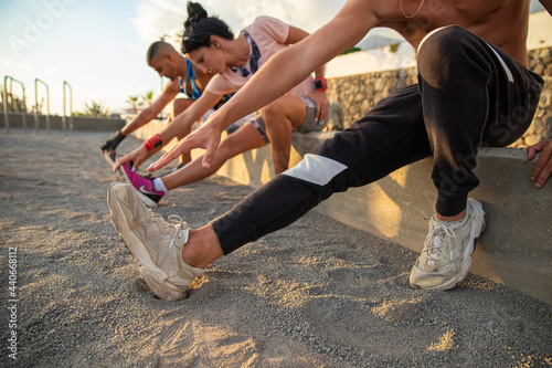 Fotografie A group of three people, two men and a woman, do some stretching exercises, they are sitting and touch their feet with their hand, warm-up pre work out in the open air in an outdoor gym