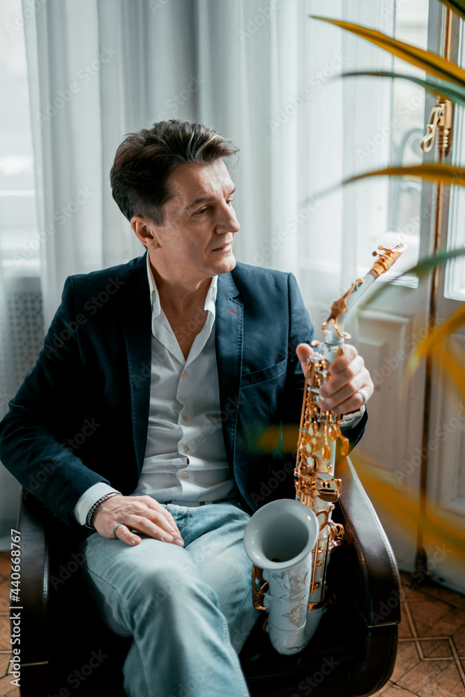 Obraz premium An adult gray-haired male musician artist sits at home and holds a saxophone in his hands.