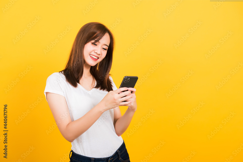 Asian happy portrait beautiful cute young woman teen smile standing playing game or writing message on smartphone looking to the phone isolated, studio shot on yellow background with copy space Asian happy portrait beautiful cute young woman teen smile standing playing game or writing message on smartphone looking to the phone isolated, studio shot on yellow background with copy space