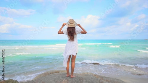 A young woman in a white sundress and straw sun hat stands on a boulder overlooking the incoming surf. photo