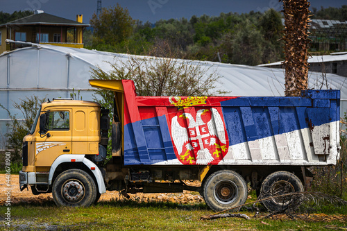 Dump truck with the image of the national flag of Serbia is parked against the background of the countryside. The concept of export-import, transportation, national delivery of goods