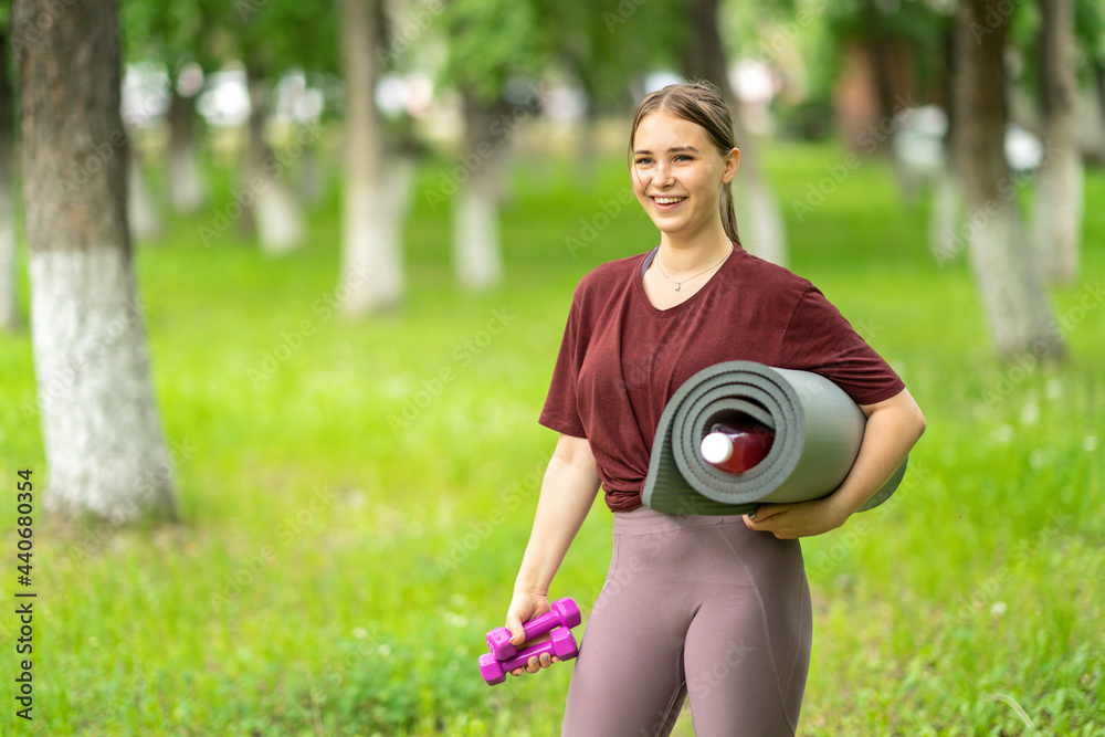 a girl with a gymnastic mat and dumbbells in her hands goes to workout in the park a girl with a gymnastic mat and dumbbells in her hands goes to workout in the park