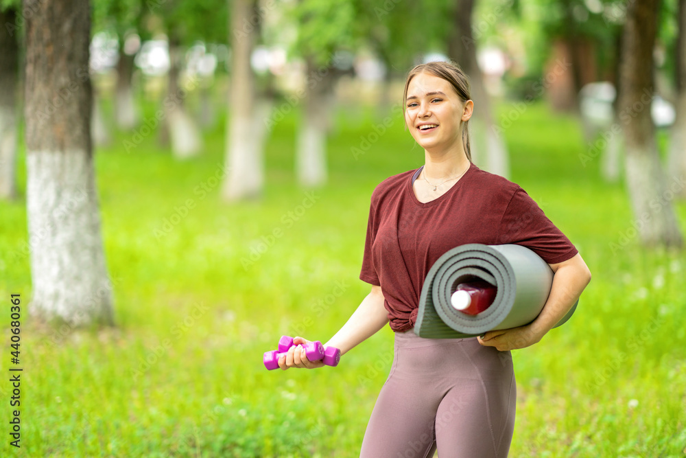 a girl with a gymnastic mat and dumbbells in her hands goes to workout in the park a girl with a gymnastic mat and dumbbells in her hands goes to workout in the park