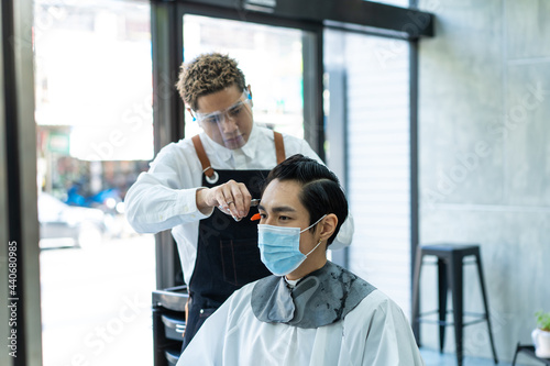 Asian hairdresser wear mask, cut young man customer's hair in salon.