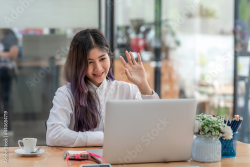 Image of a happy Asian woman waving her hand to her friend. Video call using laptop at office.