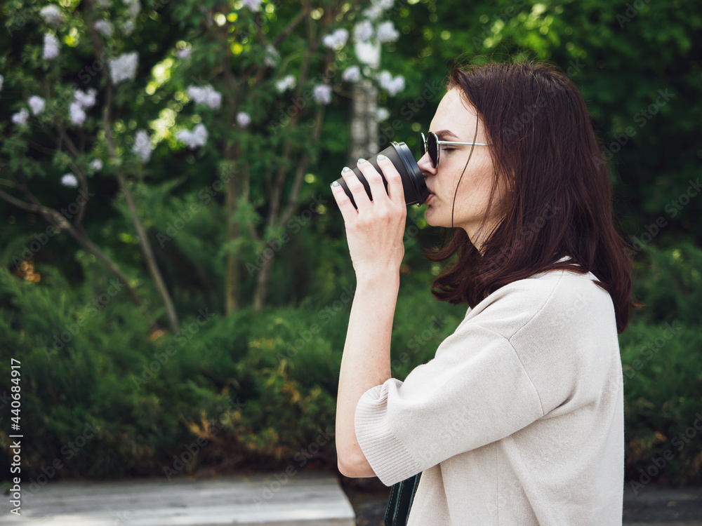 A young girl, brown-haired, with dark glasses, drinks coffee from a disposable cup, on the street. A woman drinks tea with pleasure, during a break, in the park