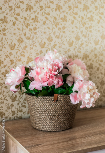 Fototapeta Naklejka Na Ścianę i Meble -  Bouquet of pink peonies in a homemade jute basket on a wooden chest of drawers in the interior of the apartment