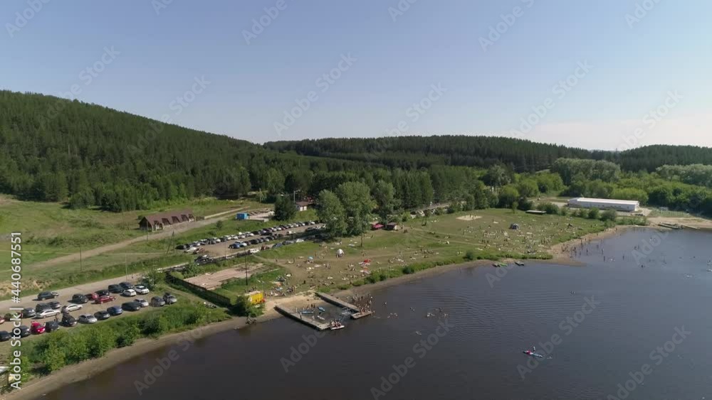 Aerial view of city beach on the outskirts of a small provincial town. People lie on the grass, swim in a pond, relax, dive, ride a jetski. Near forest, mountain and parking with many cars. Summer sun Aerial view of city beach on the outskirts of a small provincial town. People lie on the grass, swim in a pond, relax, dive, ride a jetski. Near forest, mountain and parking with many cars. Summer sun