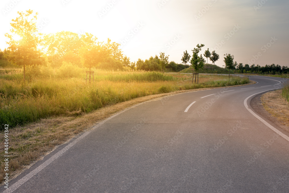 Winding road in the sunset light on the side of the road green grass. Paved village road on a summer evening. Winding road in the sunset light on the side of the road green grass. Paved village road on a summer evening.