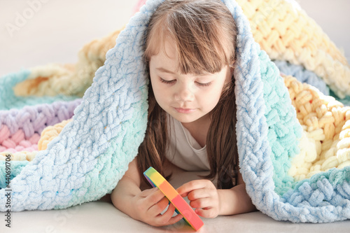 a little smiling girl hides under a rainbow blanket and plays with a multi-colored plastic toy pop it. playing at home during summer holidays