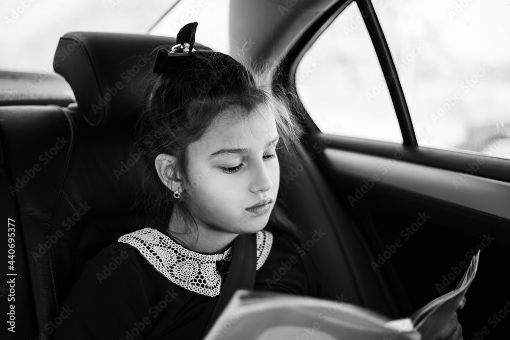Black and white photo of teenage girl reading a book inside car.