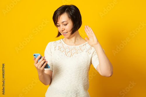 Pretty positive woman holds modern mobile phone and makes video call waves palm in hello gesture wears casual jumper isolated over yellow background. People modern technology concept. © Eugene