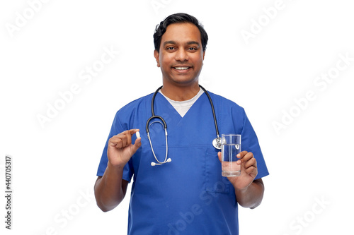 medicine, profession and healthcare concept - happy smiling indian doctor or male nurse in blue uniform with pill and glass of water over white background