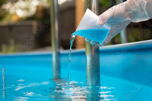 Hand holding and pouring algicide blue liquid from glass into water of swimming pool. Water purification and prevention of appearance of algae and microorganisms. photo