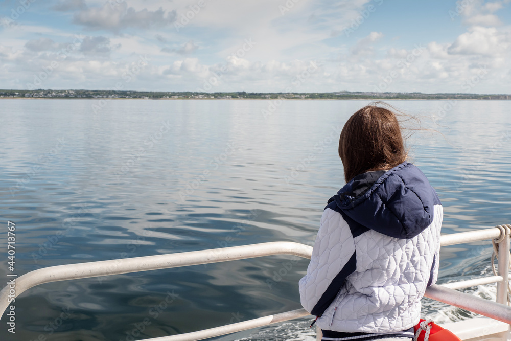 Fototapeta premium Teenager girl looking at the ocean from a deck of a ferry ship. Galway bay, Ireland. The model back to viewer. Blue cloudy sky. Girl with long hair on ocean voyage enjoys scenery. Travel concept