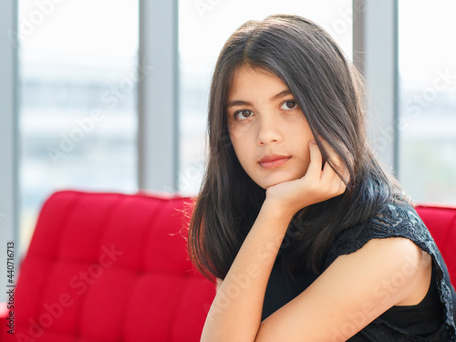 Close-up portrait shot of junior young Thai-Turkish teenager wearing trendy casual clothes sitting on the red couch and looking through the windows to outside. Young girl eyes looking sad and lonely