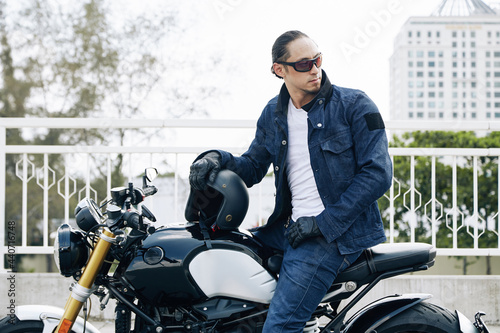 Stylish man in sunglasses, jeans and denim jacket sitting on motorcycle, taking off helmet and looking back