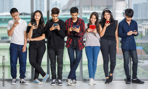 Group of seven young male and female teenagers in casual clothes standing and using their smartphones and tablet. College friends playing mobile phones. Concept of a phone addicted in adolescent