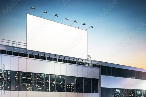Blank white billboard on business center rooftop with spotlights and night city skyscrapers reflection in building windows. 3D rendering, mockup