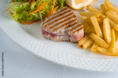 Tuna steak grilled with french fries salad of carrots, parsnips, lettuce and lemon on white plate on white background.