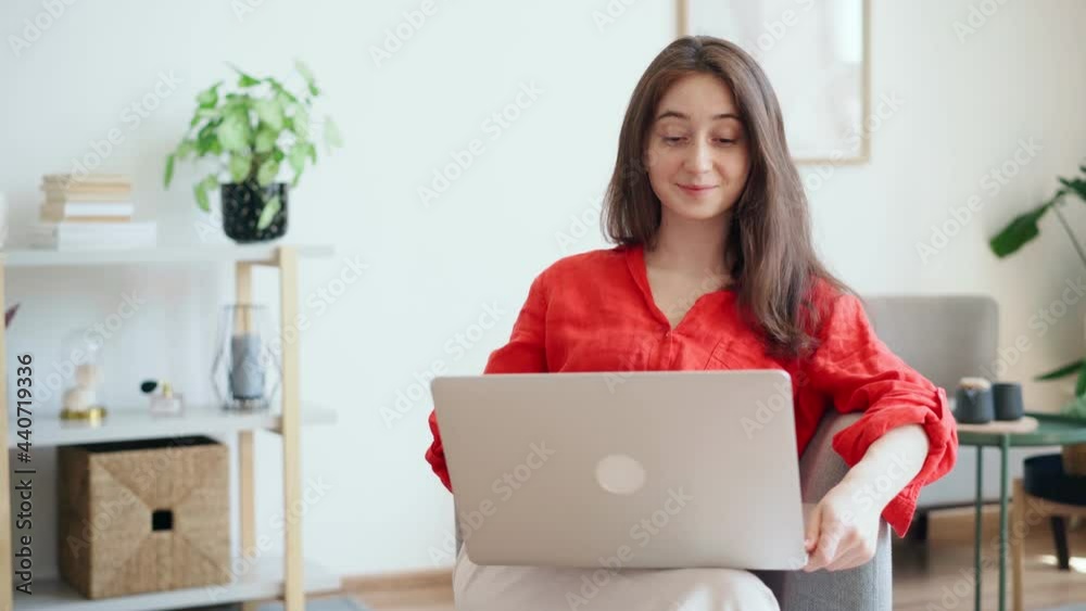 A beautiful young businesswoman in a bright red shirt is typing on a laptop while sitting in a comfortable armchair at the office A beautiful young businesswoman in a bright red shirt is typing on a laptop while sitting in a comfortable armchair at the office