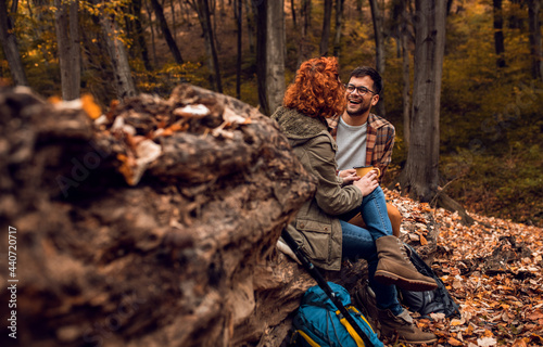 Young couple with backpack sitting on collapsed trunk resting and drinking tea after hiking in forest.