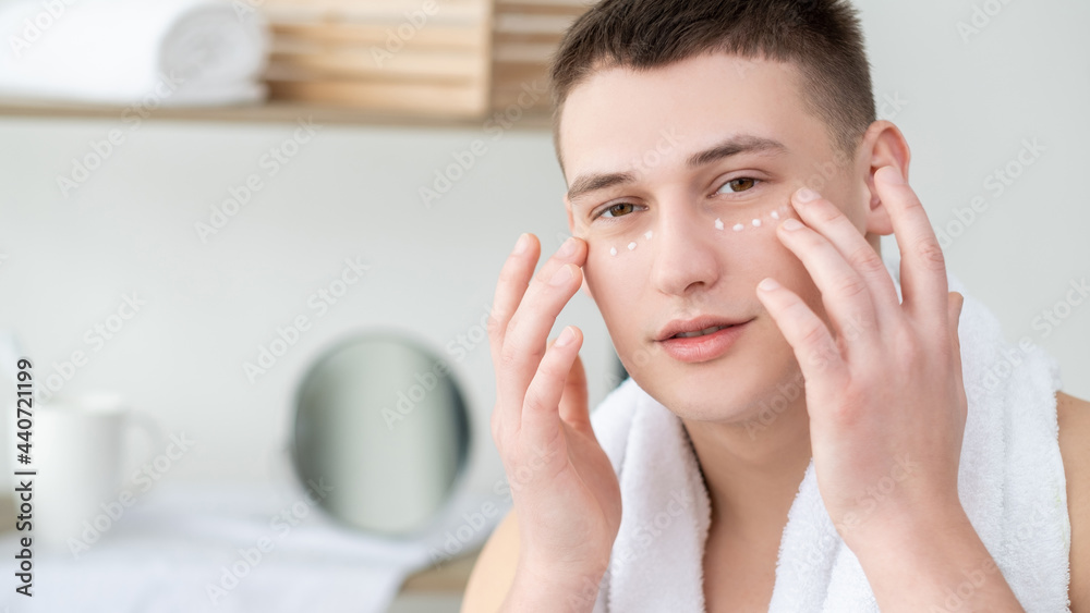 Moisturizing cream. Male skincare. Facial cosmetology. Peaceful joyful shirtless young man applying eye serum on flawless smooth clean skin on light defocused shower room background. Moisturizing cream. Male skincare. Facial cosmetology. Peaceful joyful shirtless young man applying eye serum on flawless smooth clean skin on light defocused shower room background.