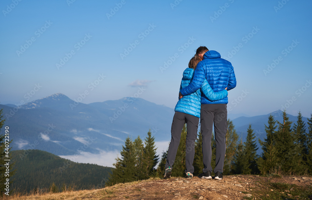 Rear view of couple in love who standing on mountain, watching around beautiful mountain scenery and tenderly embracing each other. Pleasant moment of hugs while walking in the fresh air.