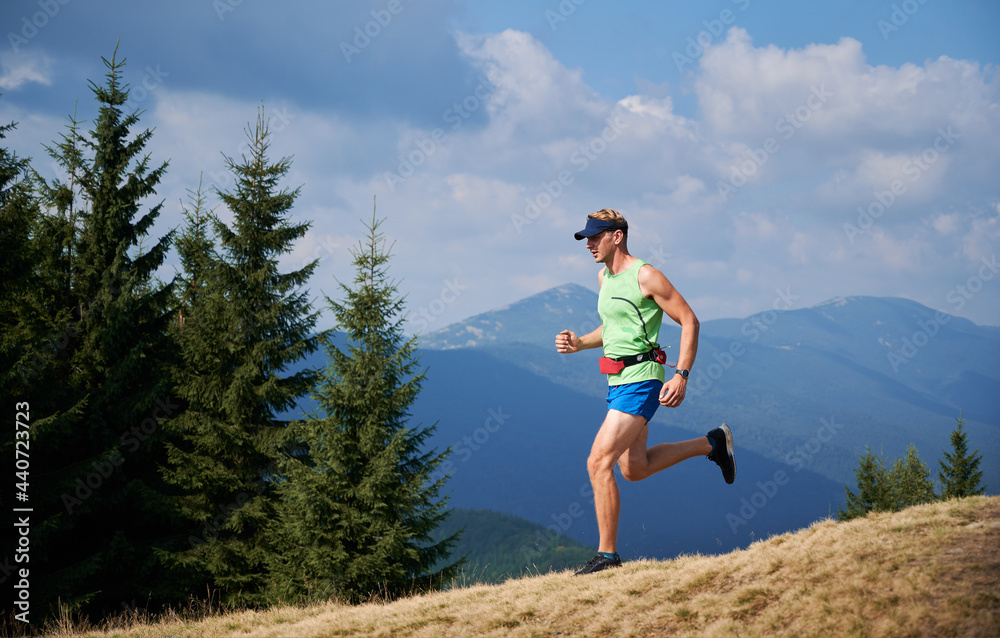 Professional young blond athlete running trail on grassy downhill of mountains in summer sunny day. Beauty mountain landscape on background.