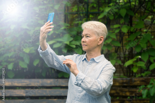 Beautiful age woman in a blue shirt with a short hairstyle  blonde takes selfies in nature in the garden in summer