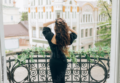 luxury woman in black evening dress stands on balcony looks at city. Back to camera rear view, turned away face. Mysterious silhouette of glamorous girl queen. Hands hold long hair, fly in wind motion photo