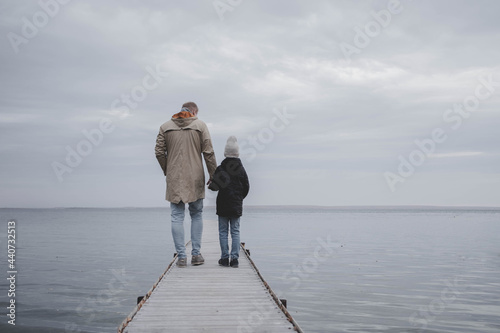 A father and son in warm jackets walk along a wooden pier over a cold lake on a cloudy winter day