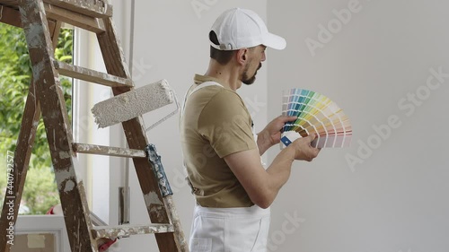 house painter man decorator choose the color using the sample swatch, work the white wall of the house to renovate, a wooden ladder, a paint roller and a green window as a background