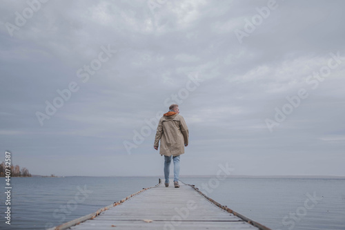 A young blond man in a light raincoat walks on a wooden pier on a lake on a foggy autumn day looking into the distance