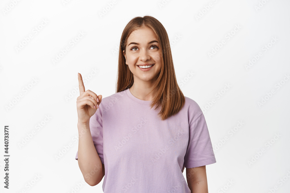 Portrait of young happy woman pointing, looking up with satisfied face expression, showing advertisement, choosing smth in store, making her decision, white background Portrait of young happy woman pointing, looking up with satisfied face expression, showing advertisement, choosing smth in store, making her decision, white background