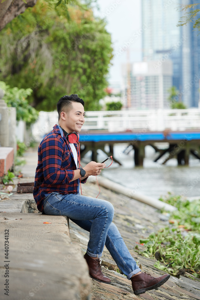 Fototapeta premium Smiling dreamy young Asian man with red headphones around neck sitting on ground near riverside and choosing track on smartphone