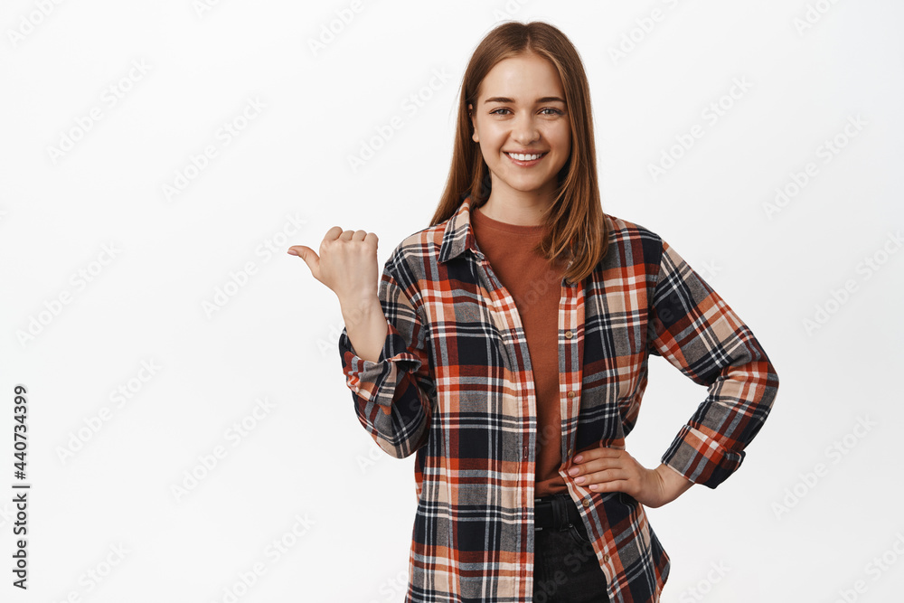 Image of smiling confident girl pointing thumb left, showing advertisement aside, announcing, indicating at chart or diagram, standing in casual clothes against white background Image of smiling confident girl pointing thumb left, showing advertisement aside, announcing, indicating at chart or diagram, standing in casual clothes against white background