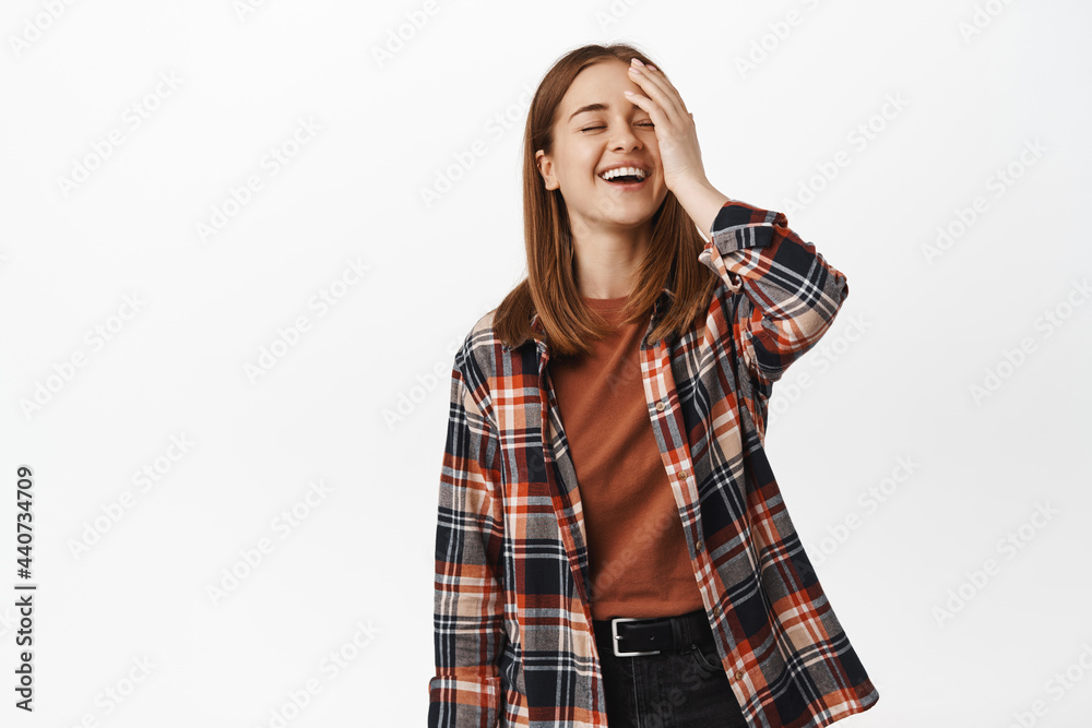 Image of young blond girl touch her face, laughing and smiling, happy face expression, carefree pose, standing in casual checked shirt and black jeans, white background Image of young blond girl touch her face, laughing and smiling, happy face expression, carefree pose, standing in casual checked shirt and black jeans, white background