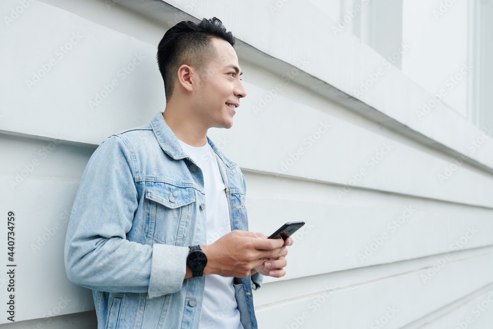 Smiling handsome young Vietnamese man in denim jacket standing with smartphone against building wall Smiling handsome young Vietnamese man in denim jacket standing with smartphone against building wall
