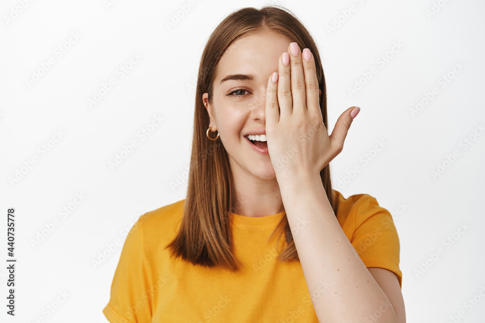 Close up portrait of smiling blond girl, cover half of face and laughing, looking at camera with one eye, checking vision in eyewear store, standing against white background Close up portrait of smiling blond girl, cover half of face and laughing, looking at camera with one eye, checking vision in eyewear store, standing against white background