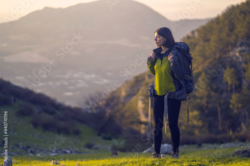 A young girl with a backpack and travel equipment looks at the amazing sunset in the mountains while she hikes along the trail. A traveler walks through the national park.