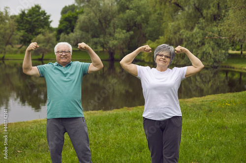 Cheerful energetic senior couple enjoy sports exercise in nature. Portrait of two happy positive healthy active old people standing in green park and flexing arms. Fitness, healthy lifestyle concept © Studio Romantic
