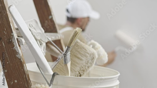 hands of house painter man decorator work of home to renovate, using roller paint and holding white bucket, a wooden ladder with paint brushes as background, close-up photo