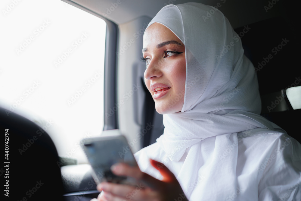 Beautiful muslim woman wearing white hijab sitting on the back seat of a car and using smartphone