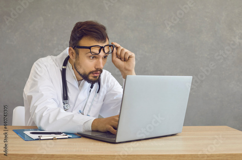 Male doctor with a shocked and confused expression looks at a laptop screen seeing a medical error. Man with frightened expression looks out from under glasses sitting at a table on a gray background