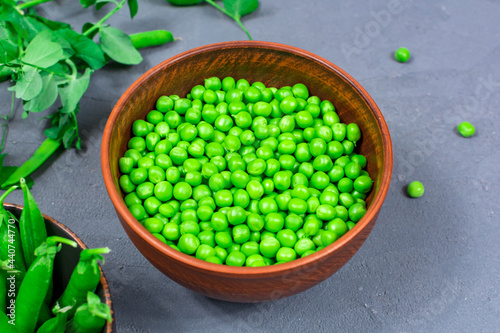 Close-up. Selective focus. Defocus. Bowls of fresh young green peas with stitches and peeled leaves on the background of shoots, sprigs of young green peas on a gray table.